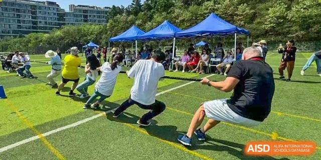 Students and adults from The British School of Nanjing participating in an outdoor activity on a grassy field with tents in the background.