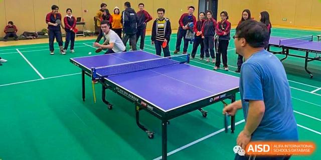 A student from The High School Affiliated To Beijing Normal University standing next to a table tennis table with a group of people in the background.