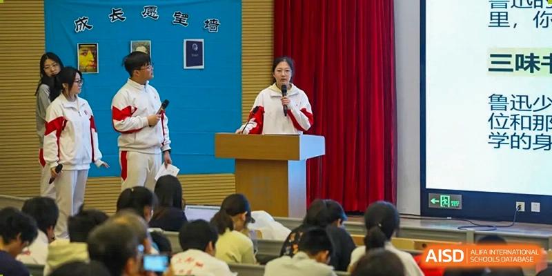 A female teacher from The High School Affiliated to Renmin University of China, Tongzhou Campus, speaks at a podium to an audience of students.