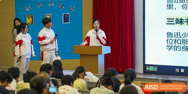 A female teacher from The High School Affiliated to Renmin University of China, Tongzhou Campus, speaks at a podium to an audience of students.