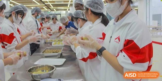 A group of students from The High School Affiliated to Renmin University of China, Tongzhou Campus, wearing aprons and gloves, prepares food in a kitchen.
