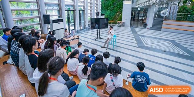 UWC Changshu China students sitting on the floor in a bright, modern space, watching a student present on a small stage.