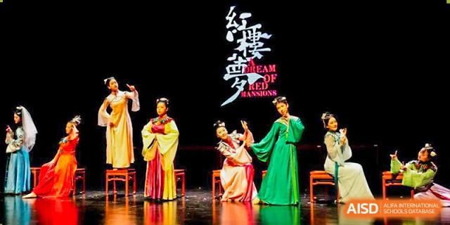 Two female dancers in traditional red and green costumes are performing on a stage with a backdrop that says "A Dream of Red Mansions" in both Chinese and English at UWC Changshu China.
