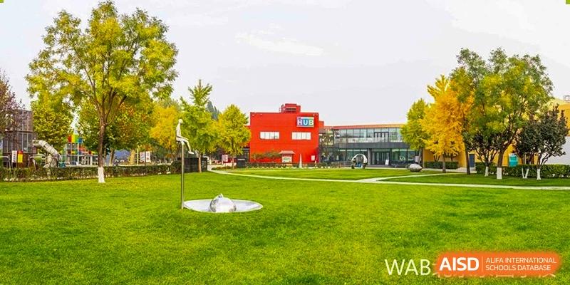 The Western Academy of Beijing campus with a large, modern red and yellow building and a green athletic field on a sunny day.