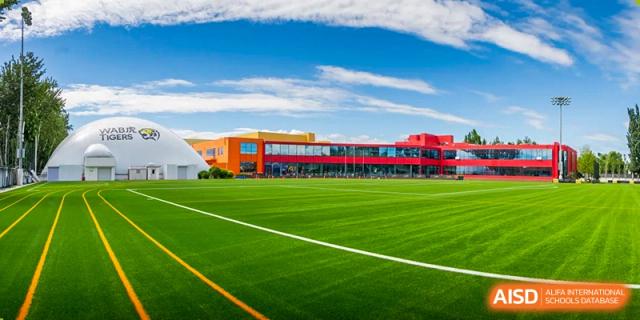 The Western Academy of Beijing campus with a large, modern red and yellow building and a green athletic field on a sunny day.