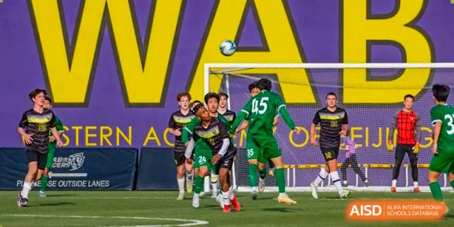 Western Academy of Beijing soccer players competing on a field with a goalkeeper defending the goal.