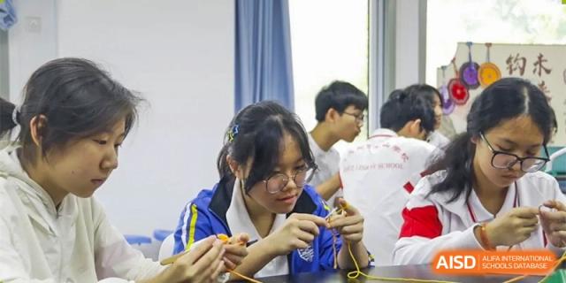 Students in white and red uniforms are learning a hands-on skill, possibly a traditional craft, in a classroom setting at the Xishan School of The High School Affiliated to Renmin University of China.
