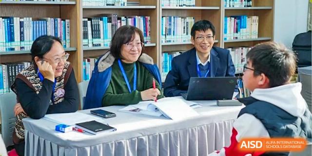 Two adults, a man and a woman, are seated at a table, possibly for a meeting or interview, with bookshelves in the background at the Xishan School of The High School Affiliated to Renmin University of China.