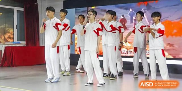 A line of male students in white uniforms with red sashes stands on a stage, possibly during a performance or ceremony at the Xishan School of The High School Affiliated to Renmin University of China.
