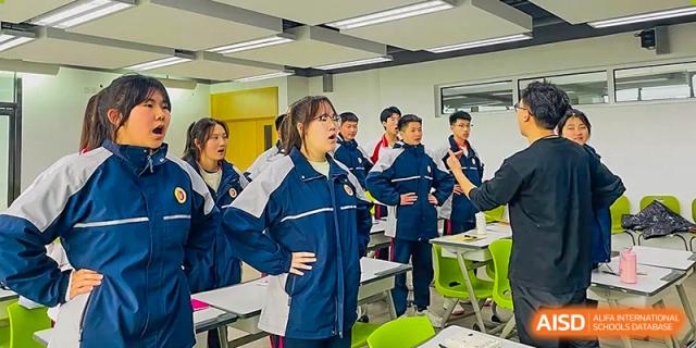 A female student leading a presentation or active discussion in a bright, modern classroom with standing students and desks at Yitian Hudson School.