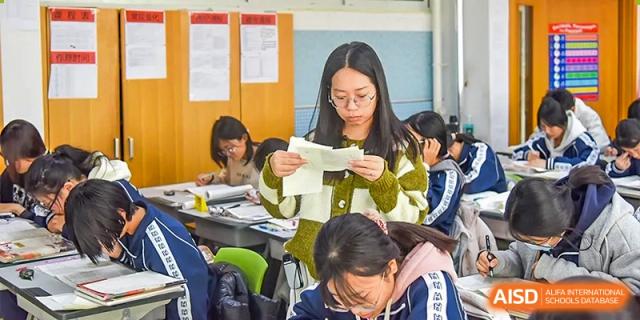 A female student reading aloud from a piece of paper during a class discussion while other students work at their desks at Yitian Hudson School.