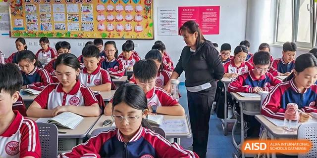 An adult teacher or instructor helping students in red and white uniforms who are sitting and working at their desks in a classroom at Yitian Hudson School.