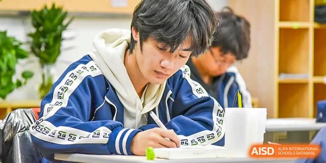 A male student in a blue and white jacket concentrating on writing notes or taking a test in a classroom at Yitian Hudson School.
