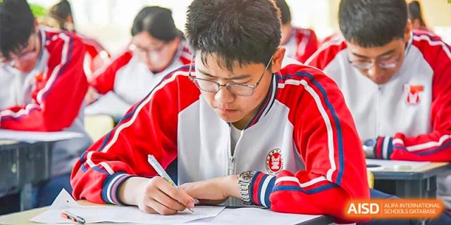 A close-up of a male student in a red and white uniform intently writing on a paper during an exam or focused study session at Yitian Hudson School.