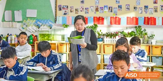 A female teacher or administrator standing and holding a clipboard while engaging with students in a bright, modern classroom at Yitian Hudson School.