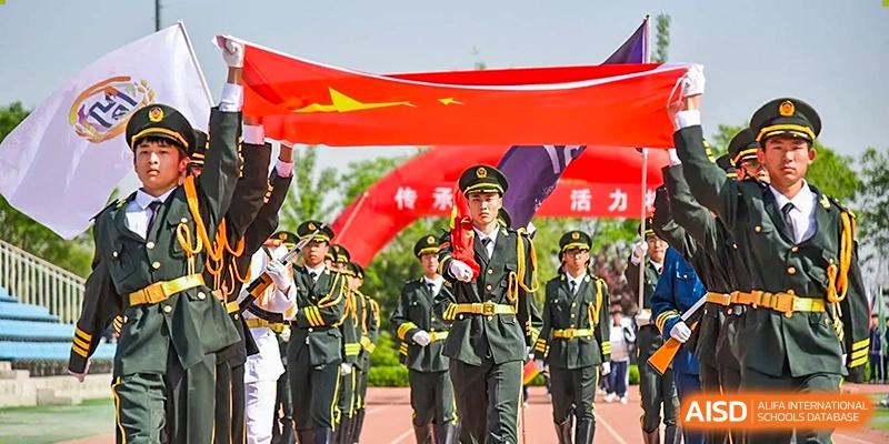 Military parade-style formation with students in black uniforms and an officer leading, holding a large Chinese flag banner at an outdoor track and field event at Yitian Hudson School.
