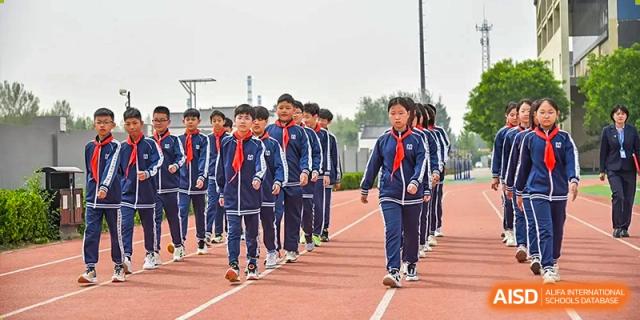 A line of students in dark blue and red scarves marching on an outdoor red track and field surface at Yitian Hudson School.