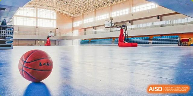A wide shot of a bright, empty indoor gymnasium with a polished wooden floor, bleachers, and a basketball hoop apparatus in the distance at Yitian Hudson School.