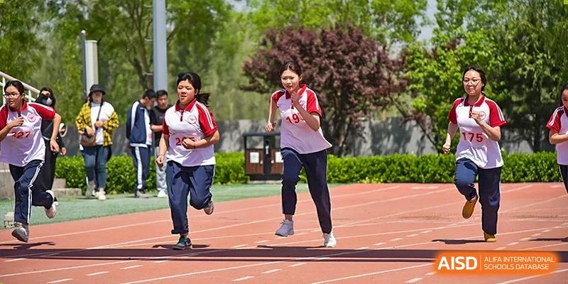 Two female students, one in a white and red shirt and one in a dark shirt, smiling while running on an outdoor red track at Yitian Hudson School.