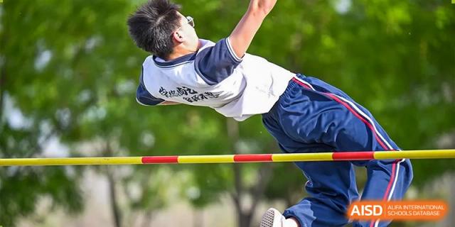 A student in a white and blue uniform successfully performing a high jump over a yellow and red bar at an outdoor track and field event at Yitian Hudson School.