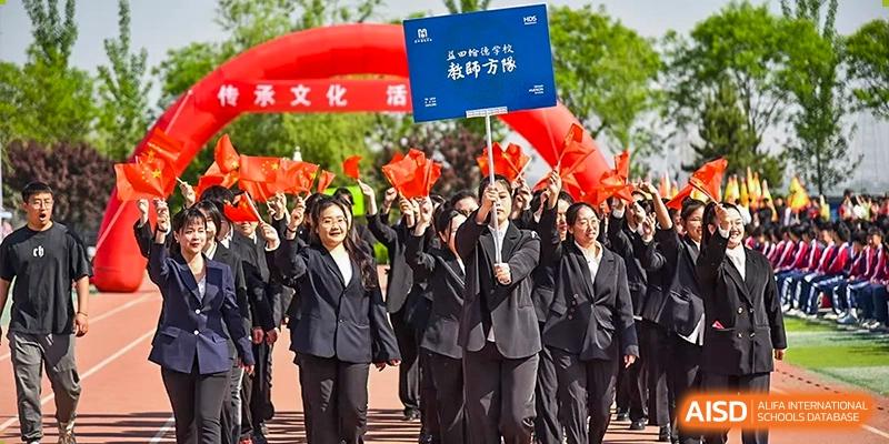 A group of cheering students and adults in black suits, raising their arms at an outdoor event with a red banner overhead at Yitian Hudson School.