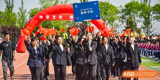 A group of cheering students and adults in black suits, raising their arms at an outdoor event with a red banner overhead at Yitian Hudson School.