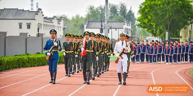 A line of students and staff in military parade uniforms marching in formation on an outdoor red track at Yitian Hudson School.