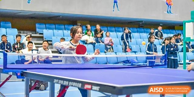 A young student playing table tennis in an arena or gymnasium with spectators seated in the background at Yitian Hudson School.