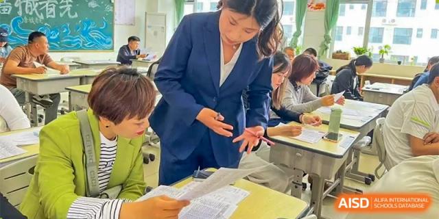 A female teacher at ZHANGQIU BILINGUAL INTERNATIONAL SCHOOL points to papers while assisting students sitting at desks during a test or class activity.