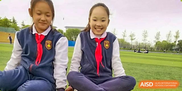 A smiling female student at ZHANGQIU BILINGUAL INTERNATIONAL SCHOOL sits cross-legged outdoors in her uniform and red scarf.