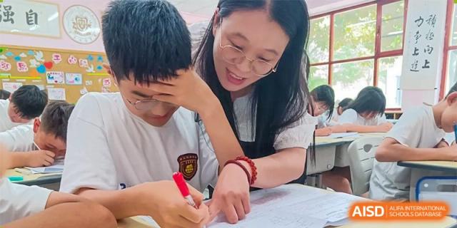 A female teacher at ZHANGQIU BILINGUAL INTERNATIONAL SCHOOL gently helps a male student with his writing or assignment.
