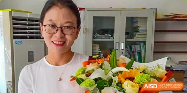 A teacher or staff member from ZHANGQIU BILINGUAL INTERNATIONAL SCHOOL smiles while holding a white and green floral arrangement indoors.