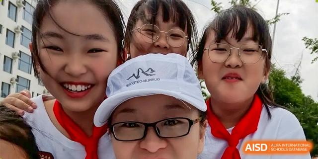 A close-up selfie of female students and a teacher from ZHANGQIU BILINGUAL INTERNATIONAL SCHOOL, one wearing a white baseball cap and glasses.