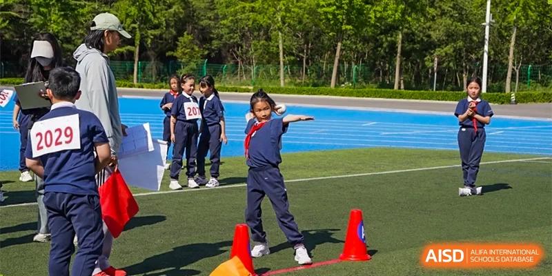 A young female student at ZHANGQIU BILINGUAL INTERNATIONAL SCHOOL performs a track and field throw event near cones on a blue track.