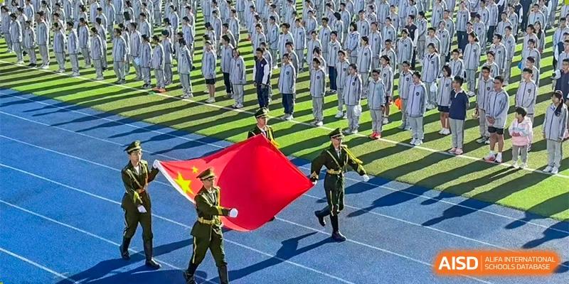 An honor guard at ZHANGQIU BILINGUAL INTERNATIONAL SCHOOL carries a large red banner past rows of students in uniform during an outdoor assembly or ceremony.