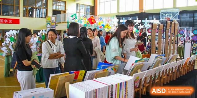 Adults and parents at ZHANGQIU BILINGUAL INTERNATIONAL SCHOOL gather around displays and books in a busy indoor hall during a school event.