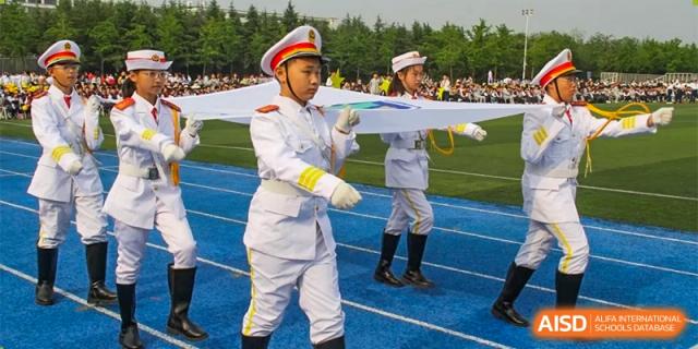 An honor guard member from ZHANGQIU BILINGUAL INTERNATIONAL SCHOOL in a white uniform holds a banner during an outdoor ceremony.