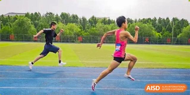 A male runner from ZHANGQIU BILINGUAL INTERNATIONAL SCHOOL sprints in a pink tank top on a blue track during a race.