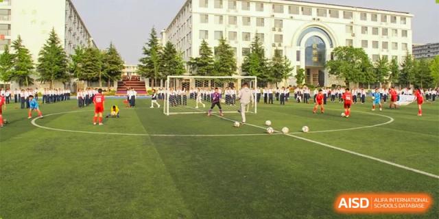 Students from ZHANGQIU BILINGUAL INTERNATIONAL SCHOOL play football (soccer) on a grass field with a goal in the background.