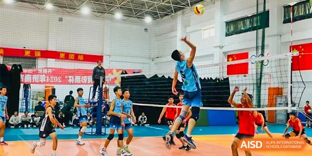 A male student from ZHANGQIU BILINGUAL INTERNATIONAL SCHOOL jumps high to spike a volleyball in an indoor gymnasium.