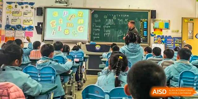A teacher from Nanjing Dongshan Foreign Language School International Department is teaching a class from a blackboard in a classroom.