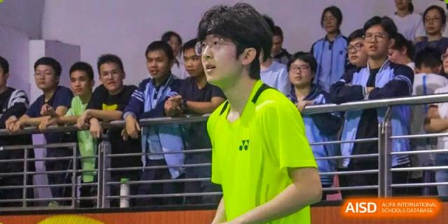 A student from Nanning No.2 High School International Division in a neon yellow shirt is playing badminton, with spectators in the background.