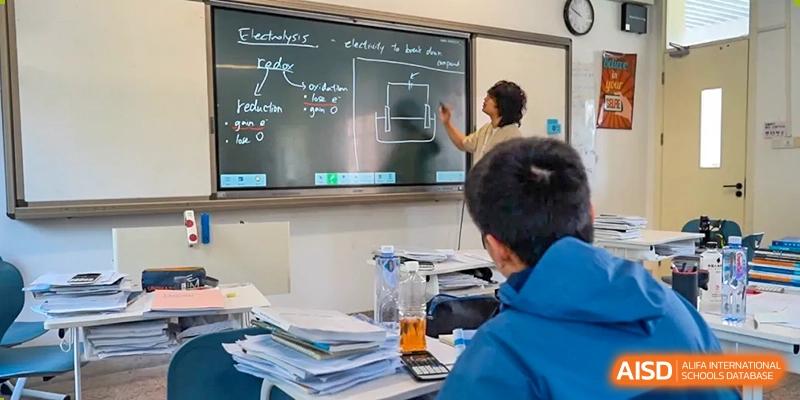 A teacher from AcaBridge Academy standing at a blackboard, explaining a concept to students seated at desks with notes and papers piled high.