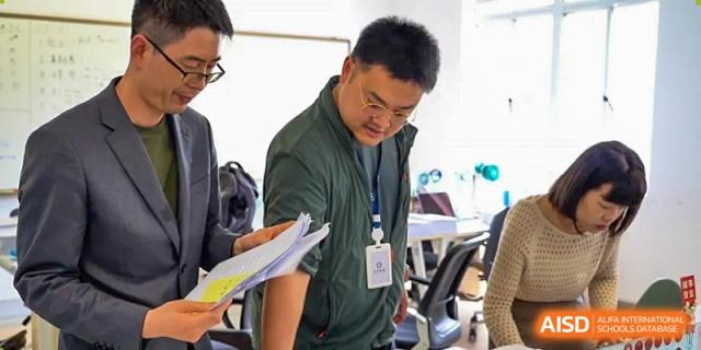 An AcaBridge Academy teacher or instructor wearing a lanyard, looking down and reviewing papers or a project binder in a classroom setting.