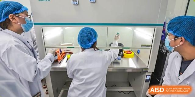 An AcaBridge Academy student or researcher, wearing a lab coat and hair net, working at a clean bench or fume hood in a science laboratory.