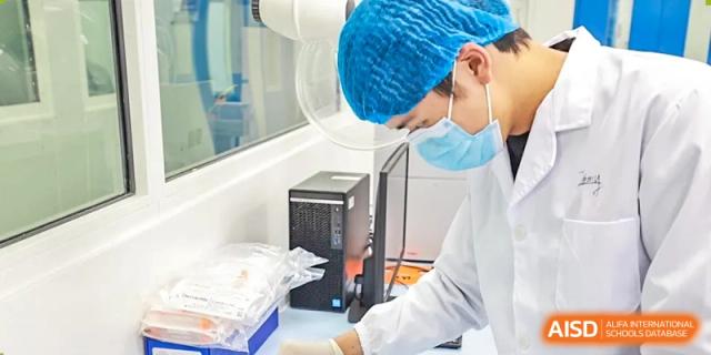 An AcaBridge Academy student or researcher wearing a lab coat, face mask, and hair net, working on a computer in a clean room or lab setting.