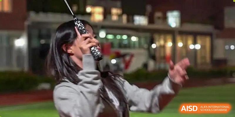 An AcaBridge Academy student holding a water bottle and cheering or singing at an outdoor nighttime event, with blurred city lights in the background.