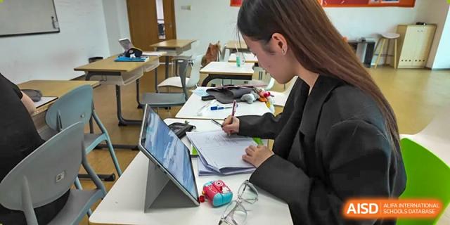 A Merchiston International School Shenzhen student focused on drawing or sketching on paper while using a digital tablet for reference or research in the classroom.