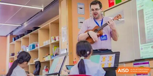 A teacher at Merchiston International School Shenzhen plays a ukulele for a student learning at a computer, integrating music into the classroom.