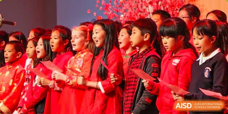 A group of young Merchiston International School Shenzhen students singing together in bright red outfits during a school performance or cultural celebration.